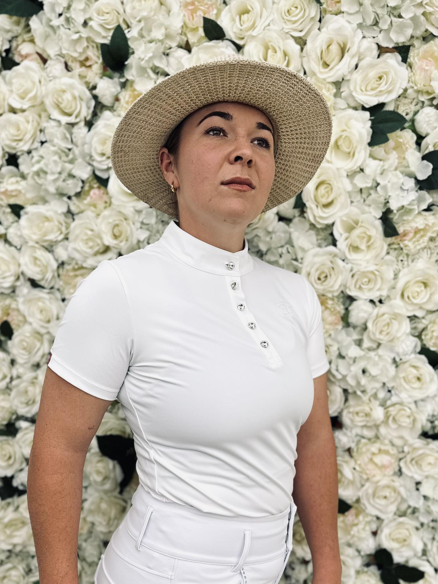 Woman in a white show shirt and straw hat with white roses backdrop, stylish for riding tights
